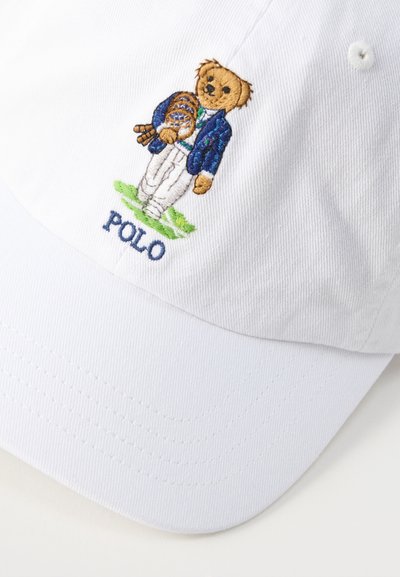 Λευκό καπέλο μπέιζμπολ με κεντητό αρκουδάκι που φορά μπλε σακάκι και άσπρο παντελόνι, κρατώντας γάντι μπέιζμπολ, πάνω από τη λέξη «POLO».