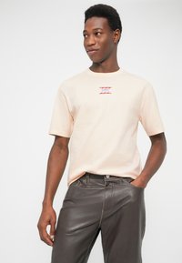 Άνδρας που φοράει ανοιχτό μπεζ μπλουζάκι με κόκκινο κείμενο και σχέδιο κυμάτων στο στήθος, σκούρα καφέ δερμάτινο παντελόνι, στέκεται με το ένα χέρι στην τσέπη.