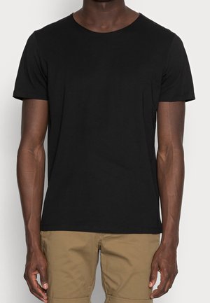 Mann in einem schlichten schwarzen T-Shirt mit kurzen Ärmeln und hellbraunen Hosen, der vor einem einfarbigen Hintergrund steht.