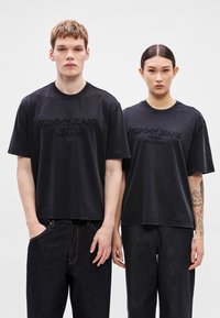 Tricouri negre din bumbac, cu croială lejeră. Prezintă logo-ul "TOMMY JEANS" în relief pe față. Asortate cu blugi din denim închis la culoare.