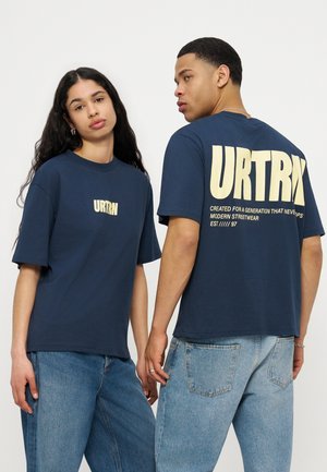 Deux mannequins portant des t-shirts URTRN bleu marine et des jeans bleus clairs, l'un faisant face à l'avant et l'autre montrant le logo et le texte au dos.