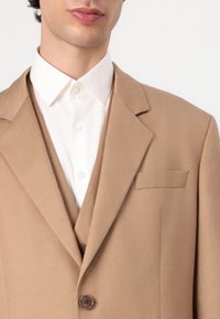 Beige blazer with a structured design, notched lapels, and a chest pocket, layered over a white button-up shirt with a collar.