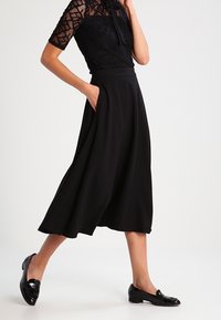 Gonna nera a trapezio con un tessuto liscio, dotata di una vita aderente e tasche laterali, abbinata a un top in pizzo nero testurizzato e mocassini.