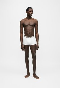 Homme mince debout pieds nus portant un boxer blanc avec une ceinture de marque comportant des logos d’ours en peluche.
