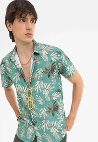 Camisa de manga corta color teal con un patrón de hojas, hecha de un tejido ligero. Presenta un cierre frontal de botones y un corte casual con cuello.
