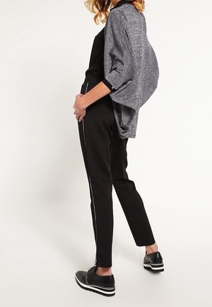 Musta istuva housut valkoisilla sivupinnoilla, yhdistettynä löysään, teksturoituun harmaaseen liiviin. Kengät ovat mustat valkoisella raidoitetulla pohjalla.