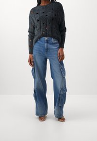 Personne portant un pantalon cargo en denim bleu à jambe large taille haute et un pull en maille gris foncé avec de grands trous, debout sur un fond blanc uni.