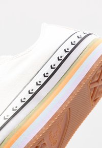 Baskets en toile blanches avec une tige texturée. Dotées d'une semelle multicolore avec des accents orange, vert, bleu et lavande, ainsi qu'un motif d'étoiles.