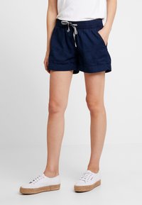 Shorts en lin bleu marine avec une taille élastique et un cordon de serrage, dotés de poches latérales et d'une coupe décontractée. Associés à des chaussures blanches.