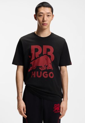 Young man wearing a black T-shirt and pants with large red "RB HUGO" logos and a stylized bull graphic on both garments.