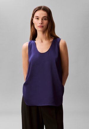 Donna con capelli castano lisci che indossa una maglia senza maniche di colore viola scuro con trama e pantaloni neri su uno sfondo grigio semplice.