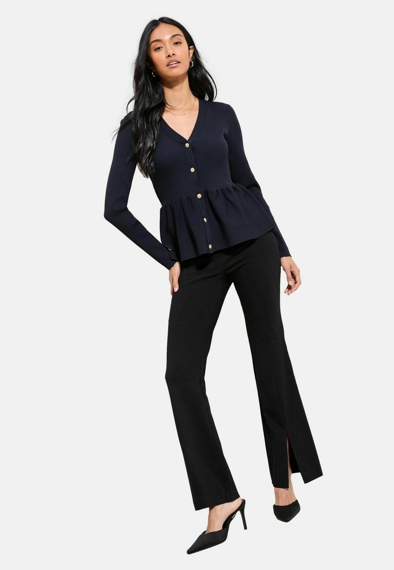Cardigan blu navy a coste con orlo a peplo, bottoni dorati e maniche lunghe abbinato a pantaloni neri con spacchi laterali e tacchi a punta.
