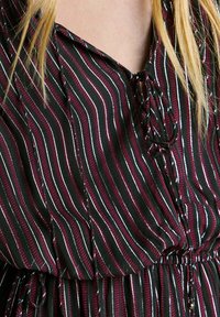 Blusa de rayas con líneas verticales en verde oscuro, burdeos y plateado. Tejido texturizado con escote en V y detalle con cordones en la parte delantera.