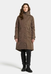 Bruine geïsoleerde parka met een hoge kraag en capuchon, met zijvakken, knoopsluitingen op de mouwen en een lengte tot de knieën. Draagt over zwarte broek en laarzen.