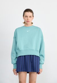 Sudadera corta verde menta con mangas largas y puños acanalados. Presenta un pequeño logo blanco de Nike en el pecho. Combinada con una falda a rayas azul marino.