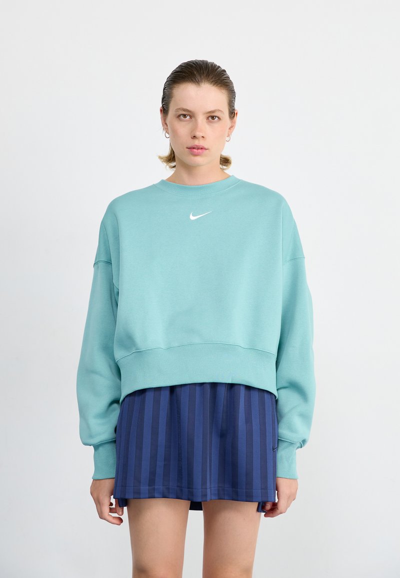 Sudadera corta verde menta con mangas largas y puños acanalados. Presenta un pequeño logo blanco de Nike en el pecho. Combinada con una falda a rayas azul marino.