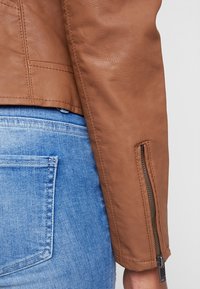 Bruine leren jas met een gestructureerde afwerking, voorzien van een ritsmanchet en verticale naaddetails, gecombineerd met blauwe denim jeans.