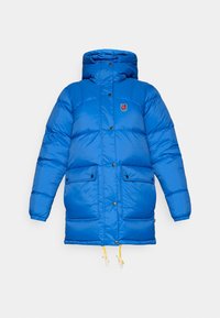 Blauwe pufferjas met een hoge kraag, sluiting met drukknopen aan de voorkant, twee grote zakken en verstelbare gele koordjes aan de onderkant.