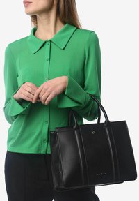 Zwarte leren tote bag met gestructureerd ontwerp, glanzende afwerking en twee handvatten aan de bovenkant. Gepaarde met een groene overhemd met lange mouwen en knopen.