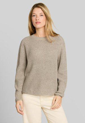 Junge Frau mit glatten blonden Haaren, die einen beigen Strickpullover und hellcremefarbene Hosen trägt, steht vor einem einfarbigen grauen Hintergrund.
