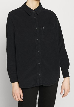 Personne portant une chemise noire à manches longues avec un petit logo blanc sur la poche poitrine, associée à un pantalon noir.