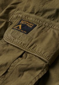 Olijfgroene utility jack met een opvallende stoffen patch met gele en blauwe branding, benadrukt de stiksels en textuur van het materiaal.