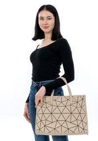 Borsa tote beige con motivo geometrico nero, design strutturato, doppi manici e superficie liscia, tenuta da una persona che indossa un top nero.