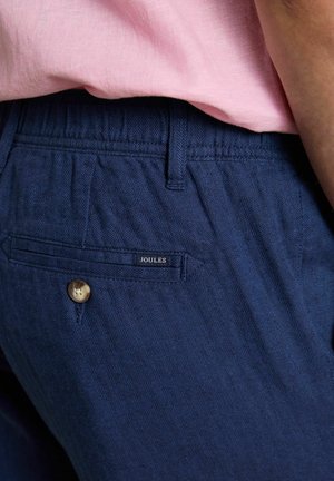 Nahaufnahme von marineblauen Fischgräten-Hosen mit einer geknöpften Gesäßtasche und einem eingesteckten pinken Hemd.