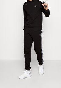 Svart sweatshirt och byxor set, med Lacoste-logotyp. Vita sidostråkar på byxorna, tillsammans med vita sportskor.