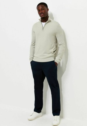 Jeune homme debout contre un mur blanc, portant un pull beige clair à fermeture éclair quart de zip, un pantalon foncé et des baskets blanches, les mains dans les poches.