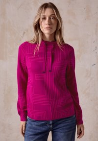 Maglione fucsia con un motivo geometrico, maniche lunghe, cappuccio regolabile con cordino e polsini a coste, abbinato a jeans blu.