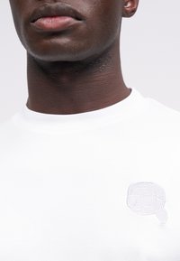 T-shirt de algodão branco com decote redondo, apresentando um desenho bordado de um rosto com óculos de sol na zona do peito esquerdo.