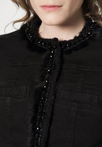 Svart denimjacka med en pälskrage prydd med svarta paljetter. Anmärkningsvärda detaljer inkluderar framfickor och texturerad trim.