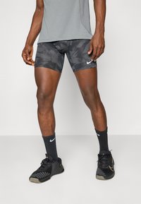 Fekete álcázott sportnadrág, testhezálló fazonnal és fehér Nike logóval, fekete bordázott zoknikkal és fekete sportcipővel párosítva.