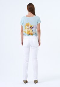 Chemise bleu clair avec imprimé floral et libellules dans le dos, associée à un pantalon cargo blanc près du corps et des sandales à talons vertes. Vue de dos.