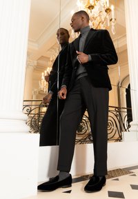 Hombre con blazer de terciopelo negro, suéter de cuello alto gris y pantalones negros, de pie junto a una pared con espejos en un interior elegante con candelabro y columnas blancas.