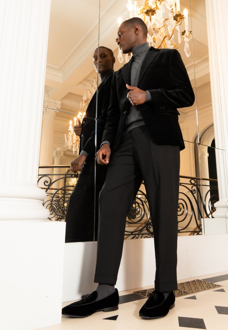 Hombre con blazer de terciopelo negro, suéter de cuello alto gris y pantalones negros, de pie junto a una pared con espejos en un interior elegante con candelabro y columnas blancas.