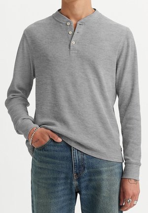 Personne portant un t-shirt henley gris côtelé à manches longues avec trois boutons, légèrement rentré dans un jean bleu, montrant des tatouages sur les mains et des bagues en argent.
