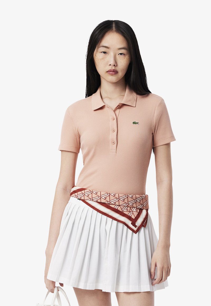 Paita polo dalam fabric reben peach lembut dengan kerah dan placket butang; dipakai bersama skirt berlipat putih dan ikat pinggang bercorak di pinggang.