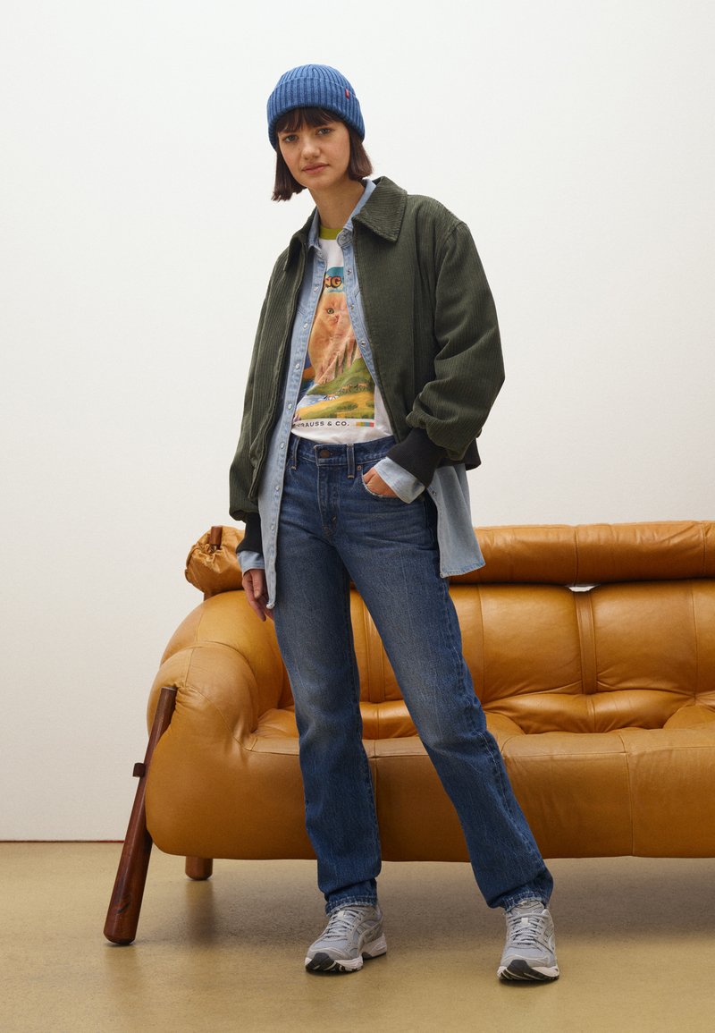 Chaqueta de pana verde sobre una camisa de mezclilla, camiseta gráfica, gorro de punto azul, jeans azules y zapatillas grises. Sofá de cuero marrón en el fondo.