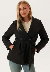 Manteau noir double boutonnage avec un large col, taille ceinturée et poches latérales, fait de tissu épais, porté sur un léger pull en maille.