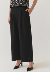 Zwarte wijde pantalons met plooien, met een gladde textuur en een gestructureerde pasvorm. De outfit wordt aangevuld met puntige zwarte hakken.