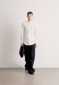 Λευκό πουκάμισο με μακριά μανίκια και όρθιο γιακά, σε συνδυασμό με σκουρόχρωμο τζιν παντελόνι. Το πρόσωπο κρατά μια μαύρη δερμάτινη τσάντα. Απλό φόντο.