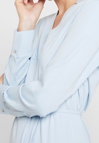 Blusa azul claro con mangas largas y ajustadas, y puños abotonados. Tejido suave con una textura lisa y un sutil detalle de drapeado.