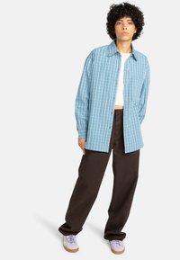 Lichtblauw geruit shirt met lange mouwen, knoopsluiting aan de voorkant en borstzak; gedragen met donkerbruine broek en paarse sneakers.