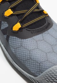 Scarpa sportiva grigia con un motivo esagonale testurizzato, dettagli neri e lacci gialli. La parte superiore è realizzata in materiale sintetico.