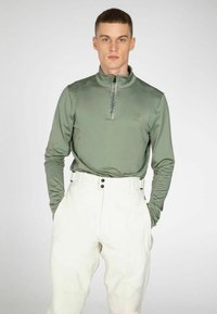 Groene lange mouwen pullover met een halflange rits en een subtiele logo op de borst, gecombineerd met hoge witte broeken met zwarte knopen.