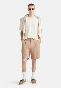 Maglione beige leggero annodato sopra una maglietta crema, pantaloni corti a quadri beige, calzini bianchi e scarpe in suede marrone. Outfit casual moderno.