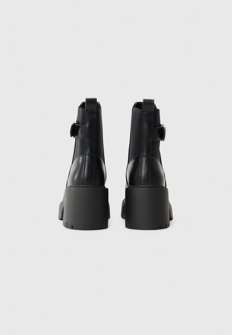 Stivali neri in pelle alla caviglia con una suola a piattaforma spessa, caratterizzati da una fascia laterale e un dettaglio con fibbia. Texture liscia con minimi accenti di cucitura.