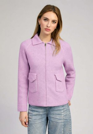 Femme aux cheveux longs portant une veste zippée violette claire avec des poches avant et un jean bleu délavé sur fond uni.